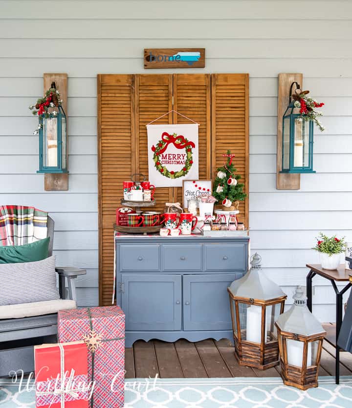 A festive porch showcases creative Christmas decoration storage ideas, featuring a blue cabinet holding holiday-themed items like a sign, small tree, and hot cocoa decor. Two lanterns rest on the floor beside a large gift box wrapped in red paper.