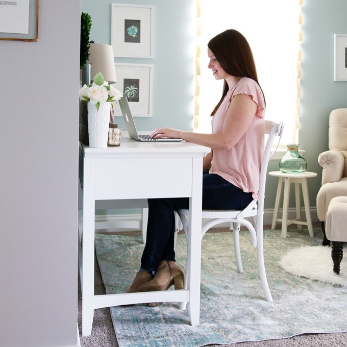 woman in pink shirt sitting at white desk looking at a laptop