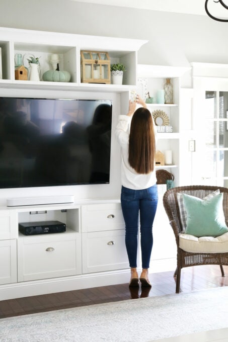 woman decorating a white entertainment unit with aqua, white, and neutral fall decor