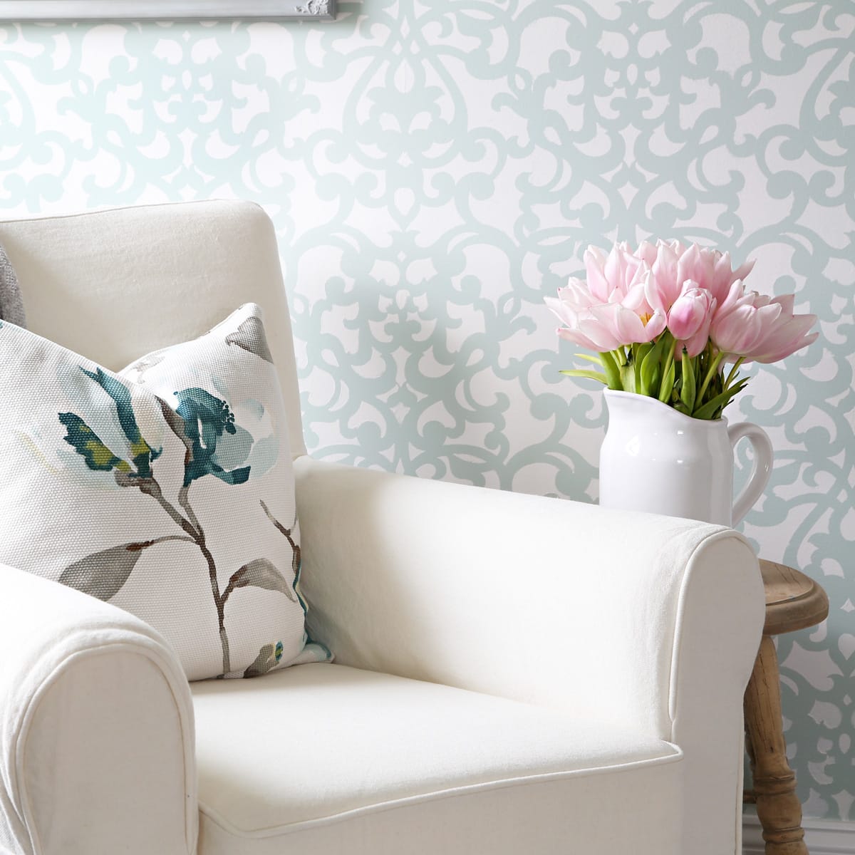 Stenciled damask wall in aqua and white behind a crock of pink tulips and a white slipcovered chair with a white and aqua floral pillow