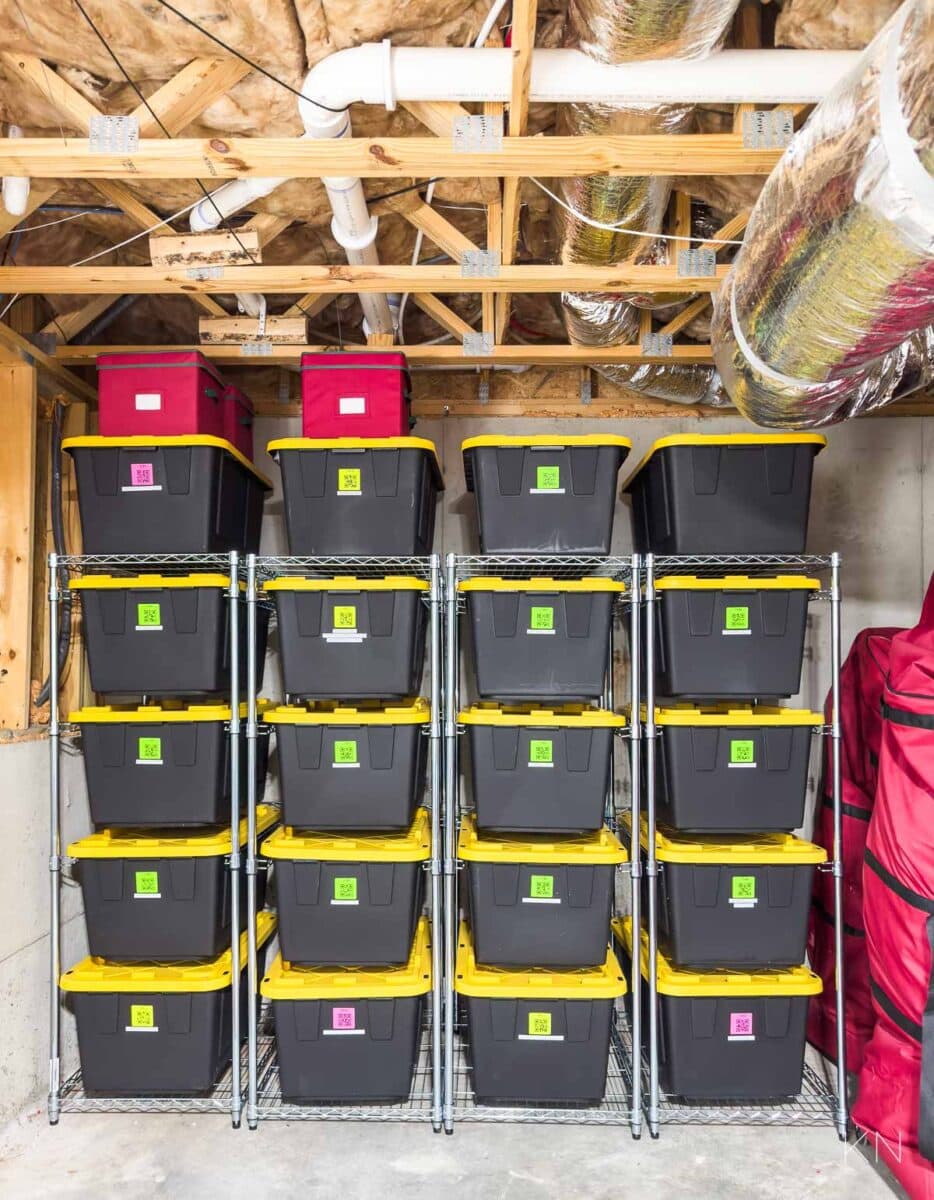 Basement storage with four metal shelves holding identical black bins with yellow lids, perfect for Christmas decoration storage ideas. Each bin has a colorful label. Two red bins are on the top-left shelf. Exposed ceiling pipes and ductwork are visible above.