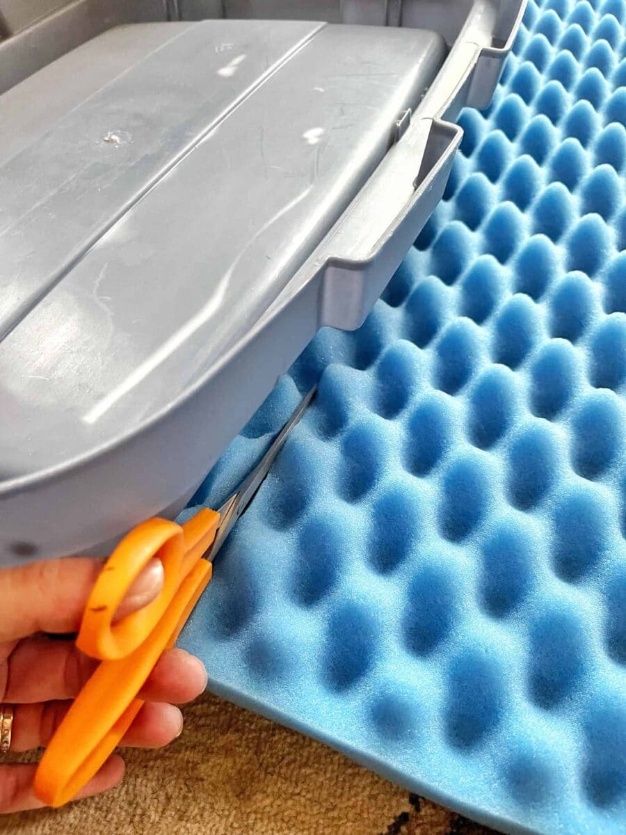 A person is using orange scissors to cut blue egg crate foam, aligning it to fit inside a gray plastic container—an efficient Christmas decoration storage idea. The foam and container sit neatly on a beige carpet, ready to protect treasured ornaments.