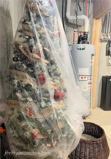 A decorated Christmas tree with red and gold ornaments is thoughtfully wrapped in plastic, embodying smart Christmas decoration storage ideas, and stored in a basement near a water heater and laundry basket.