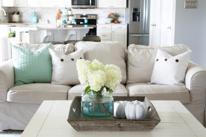 Centerpiece of white hydrangeas in an aqua jar and white pumpkins sitting in a square wooden tray on an off-white coffee table