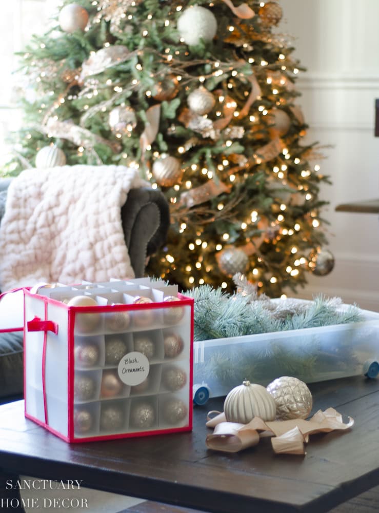 A living room showcases a festive Christmas tree adorned with lights and ornaments. In the foreground, showcasing clever Christmas decoration storage ideas, a box filled with ornaments and a container with garland are neatly placed on the coffee table while a cozy armchair with a throw blanket sits nearby.