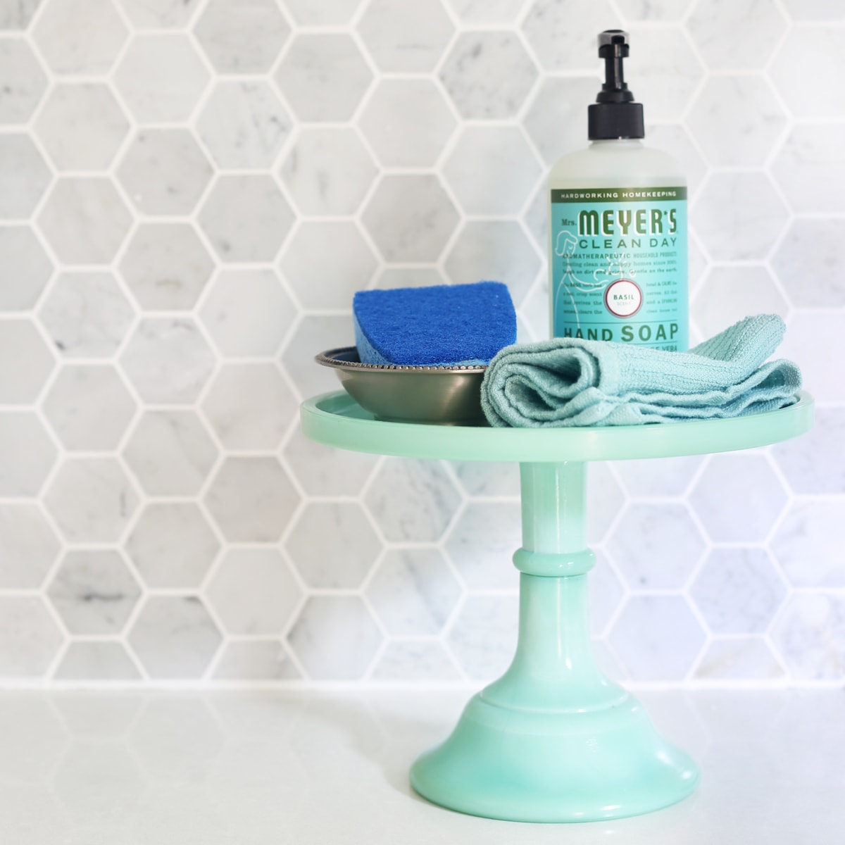 Cake Stand Holding Soap and a Sponge in Front of a Marble Hexagon Tile Backsplash