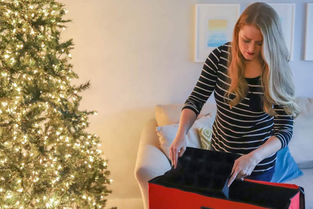 A woman with long hair in a striped shirt opens a large red storage container, showcasing clever Christmas decoration storage ideas. A decorated Christmas tree glows beside her, while framed art graces the wall behind the cozy couch.