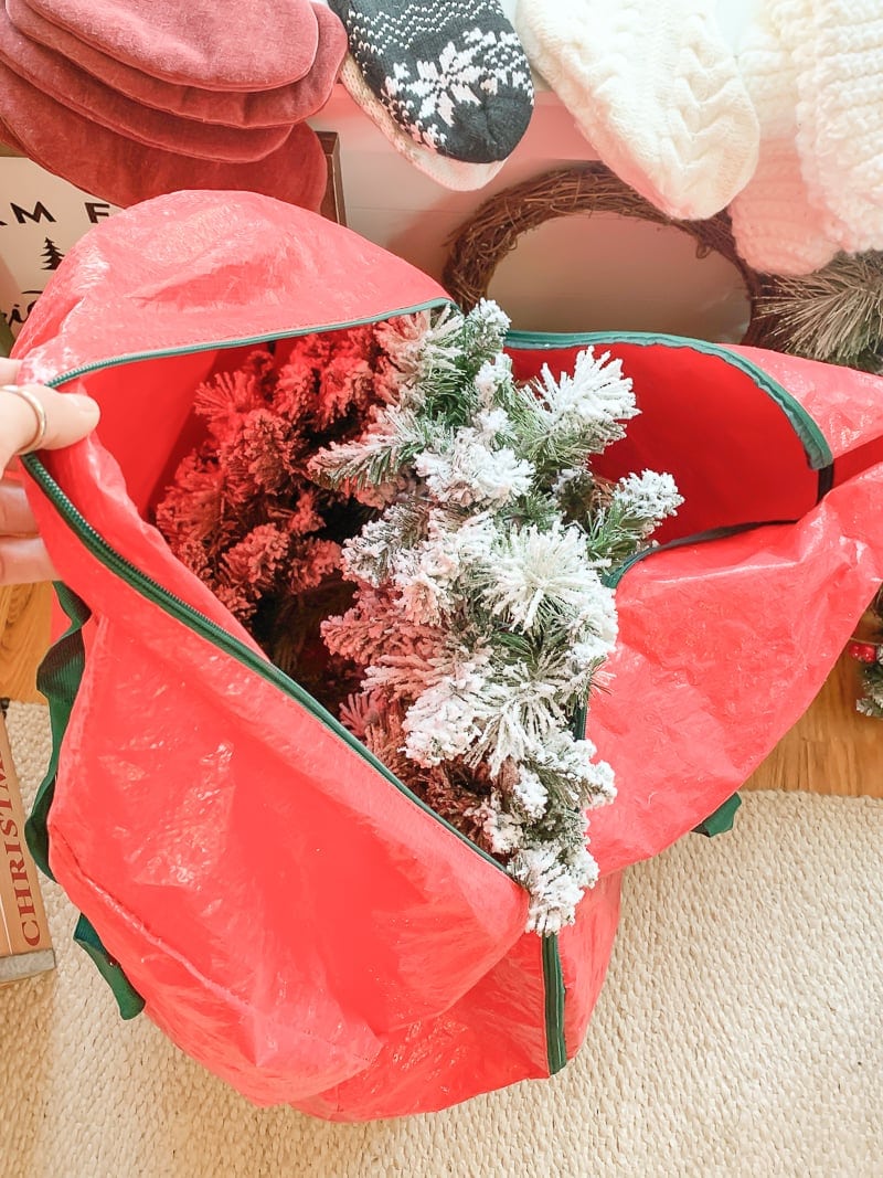 A partially opened large red storage bag reveals a frosted artificial Christmas tree, showcasing clever Christmas decoration storage ideas. The bag rests on a carpeted floor, surrounded by festive decorations, including a sign and wreaths.