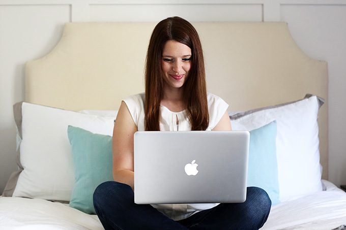 woman in a white shirt, sitting on a bed, looking at a laptop computer