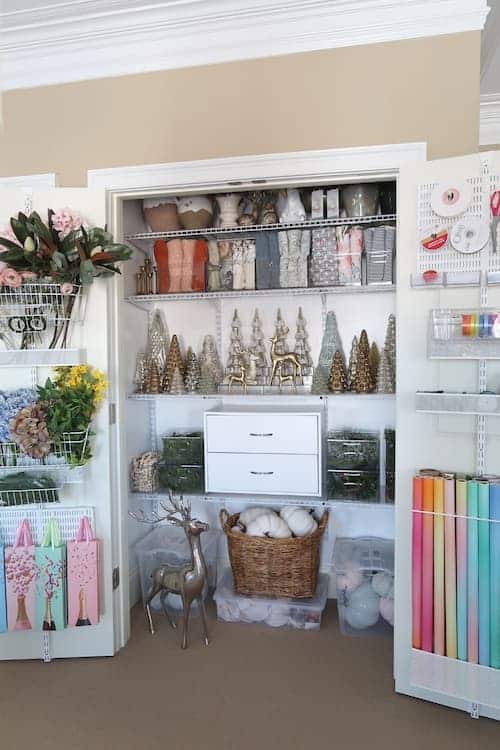 A neatly organized craft closet filled with shelves of ribbons, fabric rolls, and festive Christmas decoration storage ideas. Storage boxes and baskets hold plush toys, while decorative trees and floral arrangements add charm. The walls feature racks for cards and other crafting supplies.